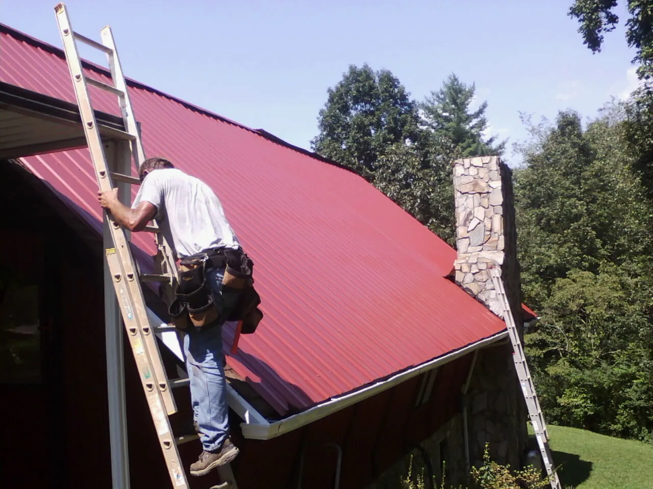 Roofer on a ladder beside a red metal roof and stone chimney — Soffit & Fascia Repair by Roofing Brothers in Crooksville